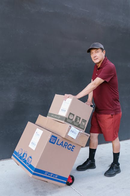 A young woman with long dark hair, wearing a white tank top, beige pants, and striped sneakers, sits cross-legged on a wooden floor surrounded by multiple large cardboard moving boxes, some sealed wit
